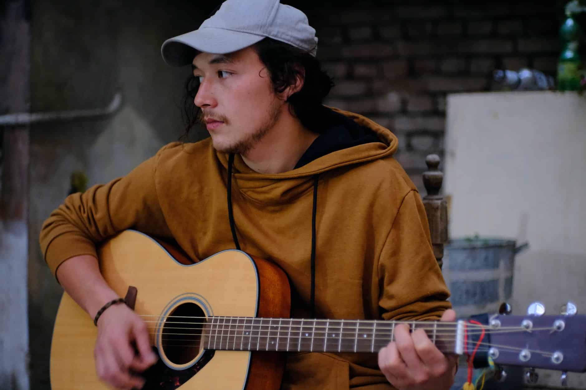 man in brown hoodie and white cap playing an acoustic guitar